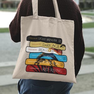 May include: A beige tote bag displaying a stack of book covers. Titles include "Light Bringer," "Dark Age," and "Red Rising." Each cover is labelled "Pierce Brown." The design incorporates a sunset and mountain graphic.