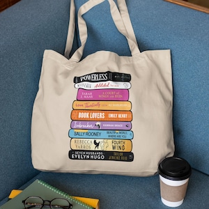 May include: Beige tote bag featuring a stack of colorful book titles, including "Powerless," "Book Lovers," and "Seven Husbands." The bag has a simple design with long handles. A coffee cup and a stack of books with glasses rest nearby.