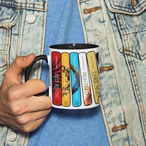 May include: A white ceramic mug with a black handle and interior. The mug features book covers with titles like "Red Rising" and "Dark Age" by Pierce Brown. The mug is held by a person wearing a denim jacket.
