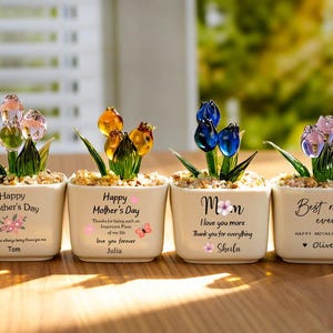 May include: Four small, square ceramic pots with glass flower sculptures. Each pot has a different message for Mother's Day, with names and floral accents. The flowers are in shades of pink, yellow, and blue.