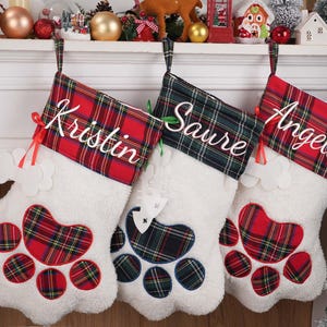May include: Three white Christmas stockings with paw-shaped designs. Each stocking has a plaid cuff with a name embroidered in white: "Kristin", "Saure", and "Angela". The paw prints are red, green, and blue plaid.
