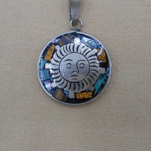 May include: A silver pendant with a sun face design. The sun is surrounded by a mosaic of turquoise, blue, and gold stones.