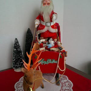 May include: A vintage Christmas display featuring Santa Claus in a red suit with a white beard, standing on a red sleigh filled with toys. A reindeer with orange antlers pulls the sleigh. The sleigh has the words "Merry Christmas".