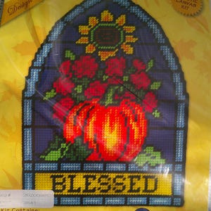 May include: A plastic canvas kit for a stained glass design. The design features a red vase filled with red roses, a yellow sunflower, and the word "BLESSED". The finished piece measures 9.5 x 13.5 inches.