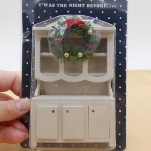 May include: White wooden dollhouse hutch with a Christmas wreath on top. The wreath is decorated with red berries and gold ornaments. The text "T'was the night before..." is printed on the packaging.
