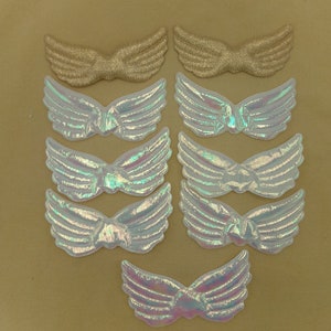 May include: A collection of nine decorative wing-shaped appliques. Two are gold-colored with a textured appearance, while the other seven are iridescent white, reflecting a rainbow of colors. The appliques are arranged on a tan background.