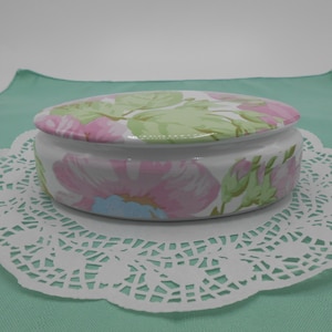 May include: Oval ceramic trinket box with a floral design in shades of pink, green, and blue. The box has a lid and is sitting on a white doily.