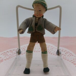 May include: A small, handmade doll dressed in traditional German clothing. The doll is wearing a green hat, a grey jacket, brown shorts, and white socks with green stripes. The doll is standing on a clear plastic stand.