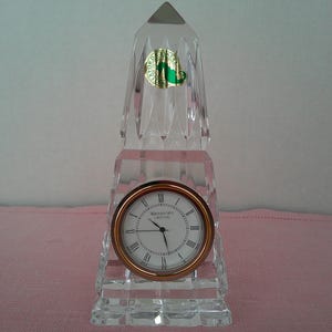 May include: A clear crystal clock with a copper-colored bezel and a white clock face with black Roman numerals. The clock is rectangular with a pointed top and a green and gold sticker that reads "Waterford Crystal".