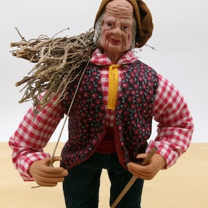 May include: A figurine of an elderly person wearing a red and white checkered shirt, a brown vest with floral print, and a brown hat. The figurine is holding a stick in one hand and carrying a bundle of twigs on their back.