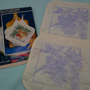 May include: Two off-white quilted pot holders with blue embroidered vegetable designs and hanging loops. A booklet titled "PAIR OF POT HOLDERS" is also present, featuring a colorful embroidered design of vegetables.