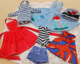 May include: A collection of doll clothes including a red dress, a blue and white striped sweater, a blue dress with embroidered flowers, a black and white striped swimsuit, a blue and red patterned shirt, and a blue denim skirt.