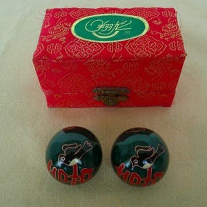 May include: A red, patterned box with a gold clasp and two dark green exercise balls. The balls have a red and gold design with a bird motif. The box has a green and gold oval logo.