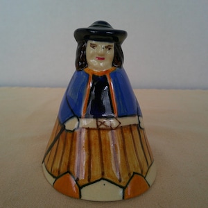 May include: A ceramic bell shaped like a figure wearing a dark blue hat, a blue cape with orange trim, and a tan skirt with vertical brown stripes. The figure has dark hair, painted facial features, and a wide base with orange accents.