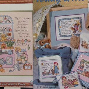 May include: A collection of cross-stitch patterns featuring teddy bears and children. The patterns include a teddy bear sitting in a rocking chair, a teddy bear holding a heart, a teddy bear with a moon and stars, and a teddy bear with a child. The patterns are all framed in blue and pink.