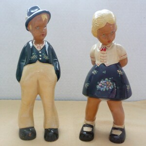 May include: Two ceramic figurines of a boy and girl dressed in traditional clothing. The boy is wearing a black hat, a blue jacket, and beige pants. The girl is wearing a white blouse with a red bow, a blue skirt with floral accents, and black shoes.
