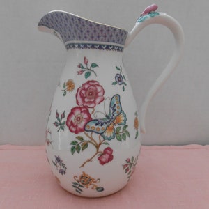May include: White ceramic pitcher with a floral and butterfly design. The pitcher has a pink butterfly with blue and yellow accents, and a pink handle with a green leaf.