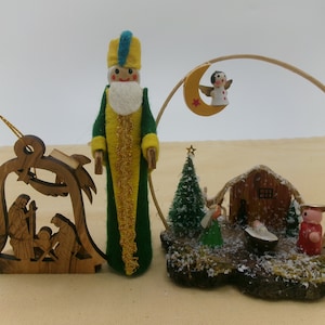 May include: A collection of Christmas ornaments. Includes a wooden bell-shaped ornament with a nativity scene, a felt figure in green and yellow, and a miniature nativity scene with a star-topped tree and angel.