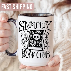 K&ouml;nnte beinhalten: Wei&szlig;er Keramikbecher mit schwarzem Henkel und Rand, mit dem Schriftzug "SMUTTY BOOK CLUB" in gotischer Schrift. Das Design umfasst ein Buch mit einem Totenkopf, Blumen und dekorativen Elementen.