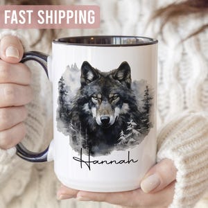 May include: White ceramic mug with a black rim and handle, featuring a watercolour image of a wolf surrounded by trees. The name "Hannah" is written in cursive below the wolf illustration. The mug is held by a person.