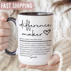 May include: White ceramic mug with a black handle and rim, featuring the words "difference maker" in a handwritten style, along with a heart symbol. The mug includes a definition and the name "Sarah".