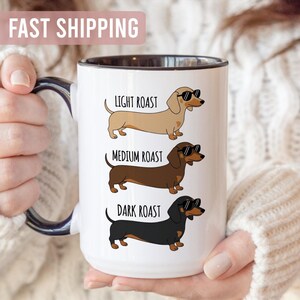 May include: White ceramic coffee mug with a black rim and handle, featuring three dachshund illustrations wearing sunglasses. The dachshunds are labeled "Light Roast," "Medium Roast," and "Dark Roast."