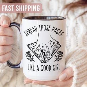 May include: White ceramic mug with a black rim and handle, featuring the text "SPREAD THOSE PAGES LIKE A GOOD GIRL" in black. The design includes a hand opening a book and rose illustrations. A cozy, warm-toned image.
