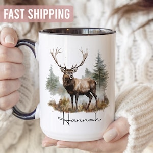 May include: White ceramic mug with a black rim and handle, featuring a watercolour illustration of a deer with large antlers in a forest setting. The name "Hannah" is written below the deer in a script font.