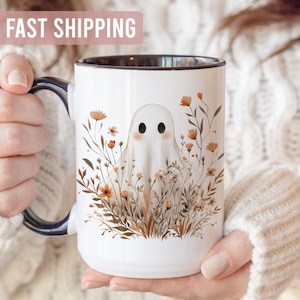 May include: White ceramic mug with a black rim and handle, featuring a whimsical ghost design surrounded by floral elements in shades of brown and orange. The mug has the text "FAST SHIPPING" at the top.
