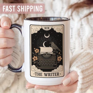 May include: White ceramic mug with a black rim and handle, featuring a tarot card design. The card depicts a vintage typewriter, a raven, and the text "THE WRITER". The background is black with a crescent moon and stars.