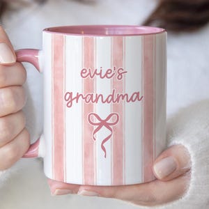 May include: A white and pink striped ceramic mug with the words "evie's grandma" and a bow graphic. The mug has a pink interior and handle. The mug is held by a person wearing a white sweater.