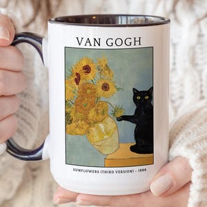 May include: A white ceramic mug with a dark handle and rim, featuring a printed graphic of a black cat sitting next to a vase of yellow sunflowers. The text VAN GOGH appears above the image, and SUNFLOWERS (THIRD VERSION) - 1888 is printed below.