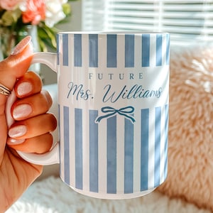 May include: A white ceramic mug featuring vertical light blue stripes. The text "FUTURE" is printed in a dark blue sans-serif font above the script text "Mrs. Williams" and a small blue bow graphic centred on the front of the mug.
