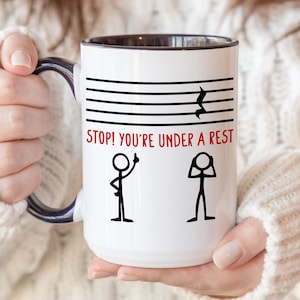 May include: White ceramic mug with a black interior and handle, featuring musical notation and the text "STOP! YOU'RE UNDER A REST" in red. The design includes stick figures. A humorous gift for musicians.