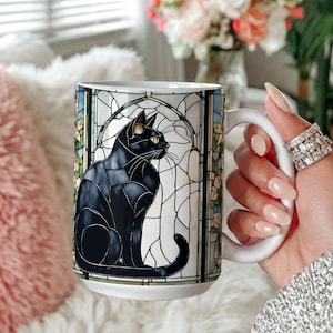 May include: White ceramic mug with a black cat design in a stained-glass style. The cat is seated, with yellow eyes and a curved tail. The mug has a large handle. The mug is held by a person.