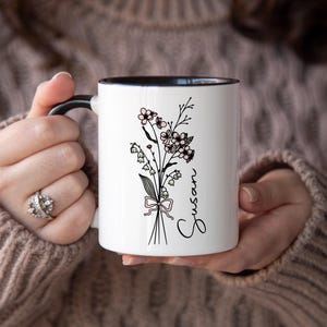 May include: White ceramic mug with a black interior and handle. The mug features a floral design with the name "Susan" in script. The flowers are black and pink, tied with a pink ribbon.