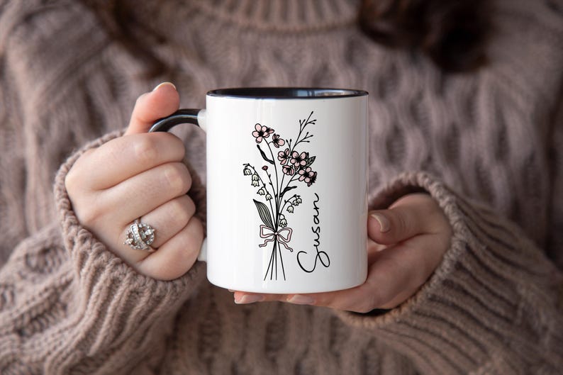 May include: White ceramic mug with a black interior and handle. The mug features a floral design with the name "Susan" in script. The flowers are black and pink, tied with a pink ribbon.