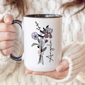 May include: A white ceramic mug with a dark interior and handle, featuring a black line art illustration of a wildflower bouquet tied with a light pink bow. The name Angela is printed in black script lettering vertically next to the flowers.