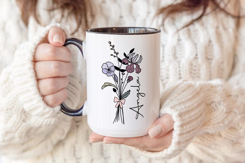 May include: A white ceramic mug with a dark interior and handle, featuring a black line art illustration of a wildflower bouquet tied with a light pink bow. The name Angela is printed in black script lettering vertically next to the flowers.