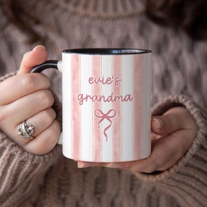 May include: White ceramic mug with pink vertical stripes and the words "evie's grandma" in a cursive font. A pink bow graphic is below the text. The mug has a black handle and interior.