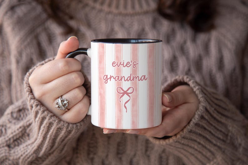 May include: White ceramic mug with pink vertical stripes and the words "evie's grandma" in a cursive font. A pink bow graphic is below the text. The mug has a black handle and interior.