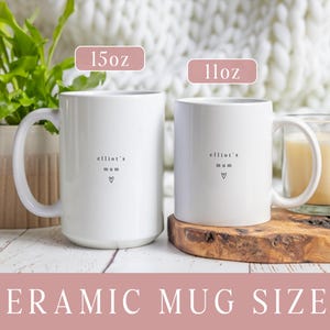 May include: Two white ceramic mugs with the text "elliot's mum" and a heart symbol. The larger mug is labeled "15oz" and the smaller mug is labeled "11oz". The mugs are on a white wooden surface. The text "CERAMIC MUG SIZES" is at the bottom.