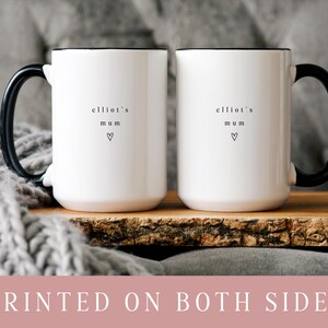 May include: Two white ceramic mugs with black handles and rims. Each mug has the text "elliot's mum" and a heart symbol printed on the front. The mugs are displayed on a wooden tray.