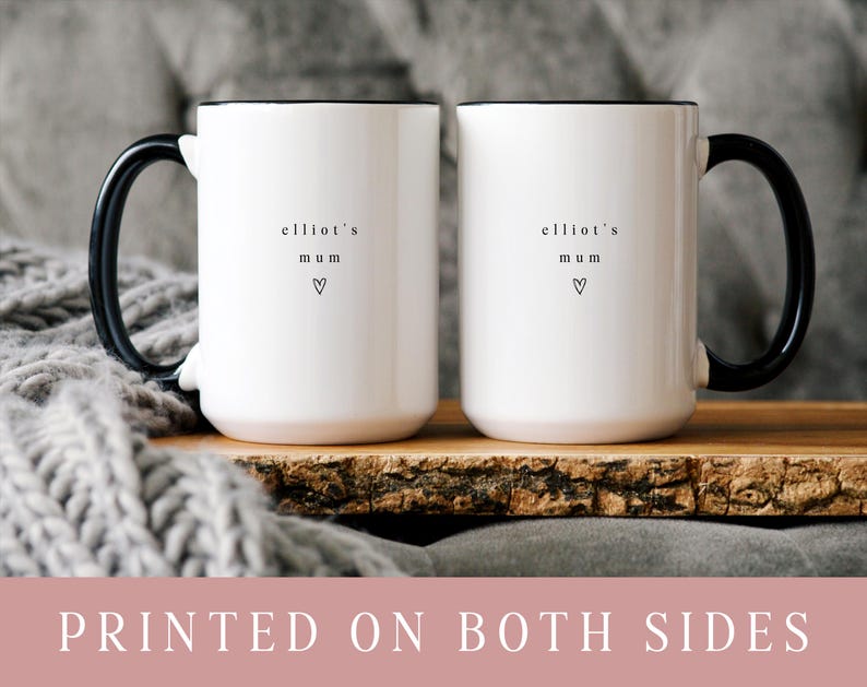 May include: Two white ceramic mugs with black handles and rims. Each mug has the text "elliot's mum" and a heart symbol printed on the front. The mugs are displayed on a wooden tray.