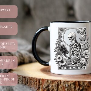 Puede incluir: Una taza de cer&aacute;mica blanca con asa y borde negros. La taza presenta una ilustraci&oacute;n en blanco y negro de un esqueleto leyendo un libro, rodeado de flores. El texto "THE READER" est&aacute; debajo.