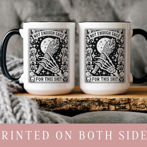 May include: Two white ceramic mugs with black handles and rims. Each mug features a black and white graphic with the text "NOT ENOUGH SAGE FOR THIS SHIT". The design includes a skeleton hand holding a bundle, a moon, and floral elements.