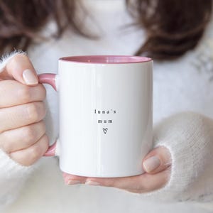 May include: White ceramic mug with a pink interior and handle. The mug has the words "luna's mum" and a heart symbol printed in black. The mug is held by a person wearing a white sweater.