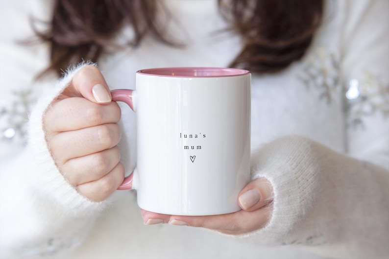 May include: White ceramic mug with a pink interior and handle. The mug has the words "luna's mum" and a heart symbol printed in black. The mug is held by a person wearing a white sweater.