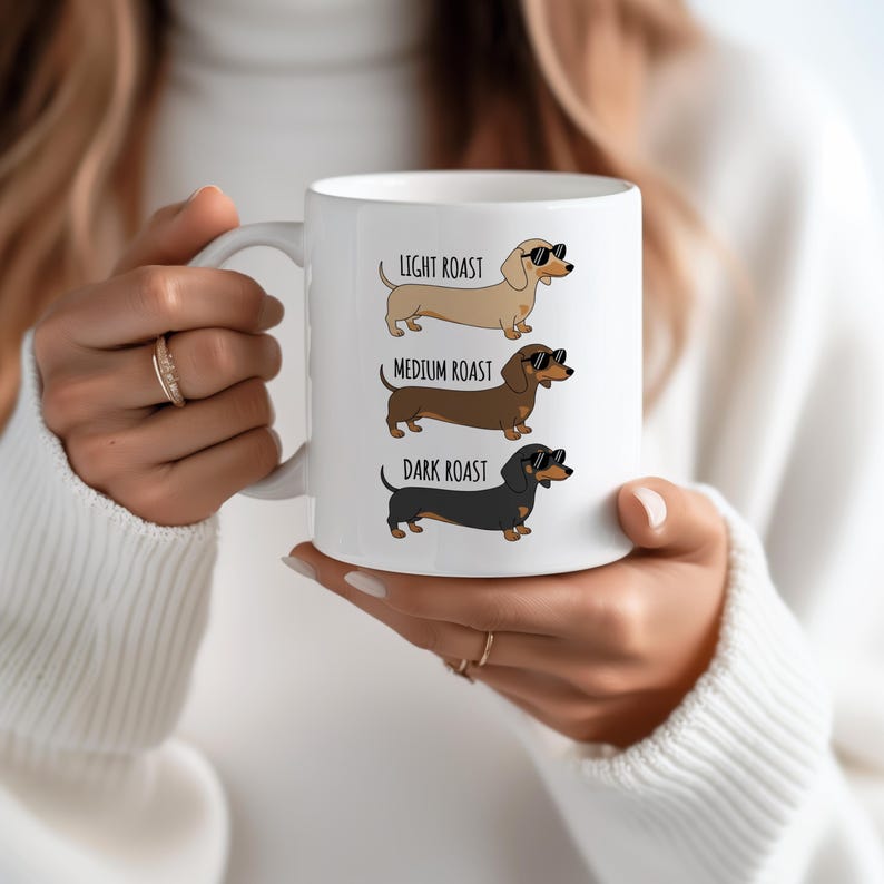 May include: White ceramic mug featuring three dachshund illustrations, each representing a different coffee roast: light, medium, and dark. The dachshunds wear sunglasses. Text above each dog reads "Light Roast", "Medium Roast", and "Dark Roast".