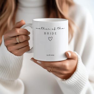 May include: White ceramic mug with the text "mother of the BRIDE" in a delicate script, a heart symbol, and the date 15.08.2025. The mug has a simple, elegant design, perfect for a wedding.